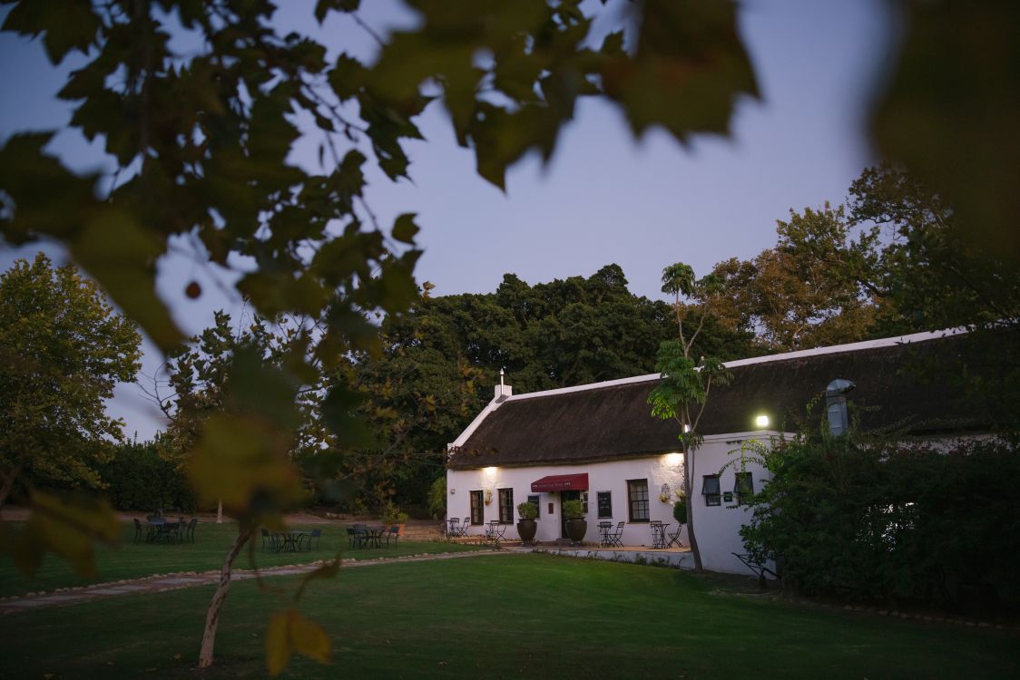 Beleuchtetes Gebäude auf dem Weingut Under Oaks in Südafrika bei Abendstimmung mit gepflegter Gartenanlage.